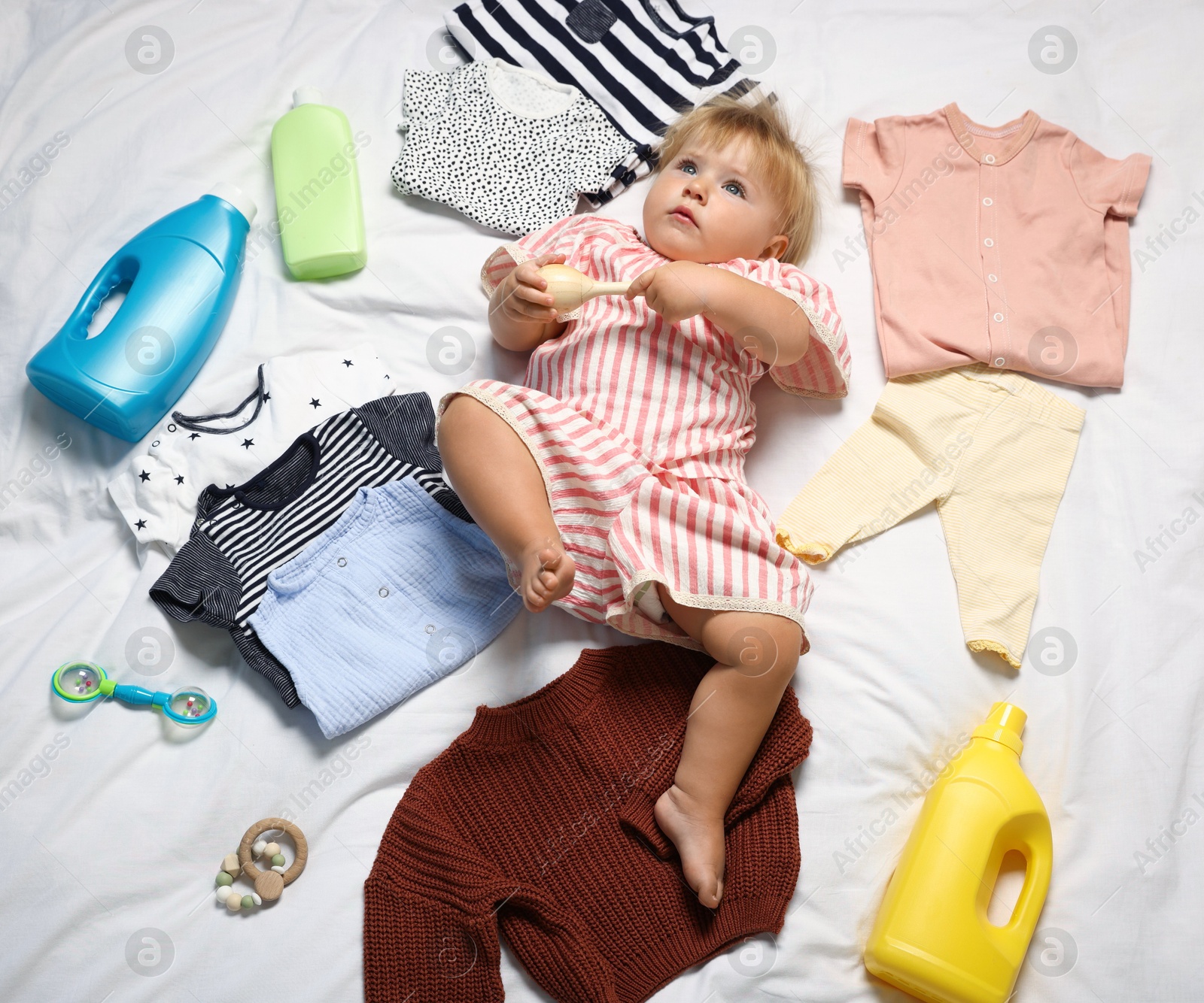 Photo of Cute baby among clean clothes and detergents on white bed linens, above view