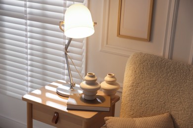 Photo of Stylish lamp, vases and book on wooden bedside table near armchair indoors