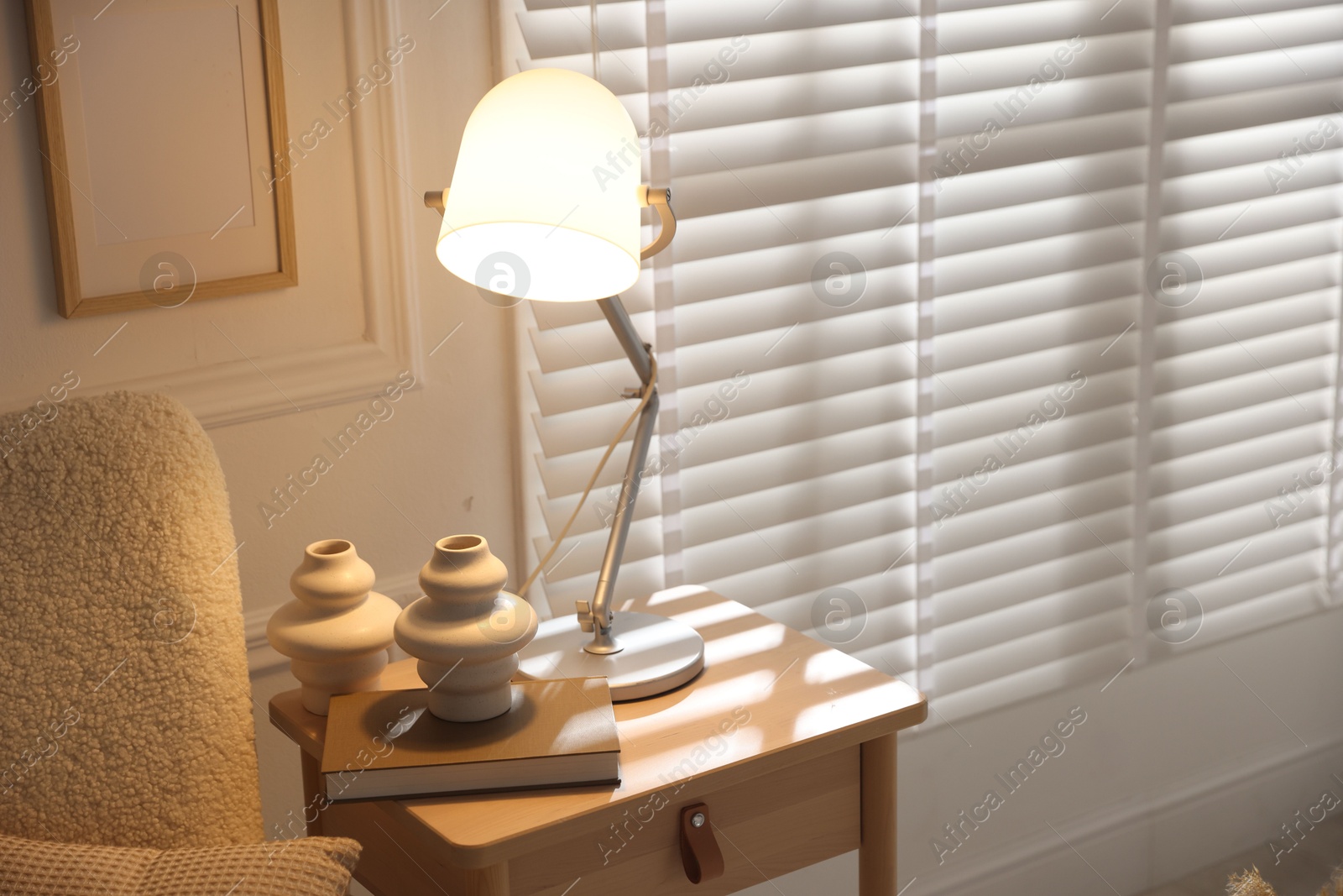Photo of Stylish lamp, vases and book on wooden bedside table near armchair indoors