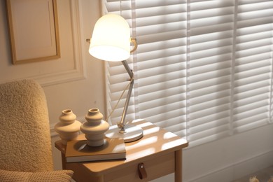 Photo of Stylish lamp, vases and book on wooden bedside table near armchair indoors