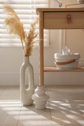 Photo of Stylish vases, dried reeds and bedside table near window indoors