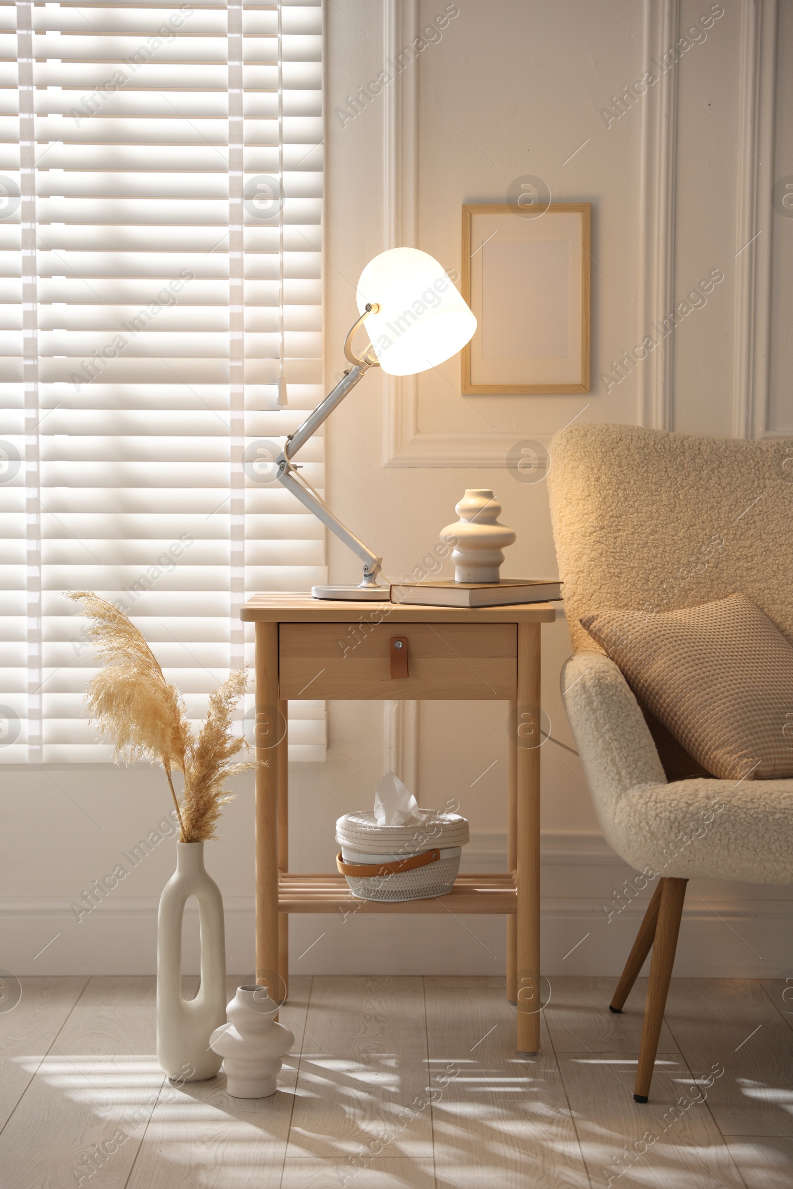 Photo of Stylish lamp, vase and book on wooden bedside table near armchair indoors