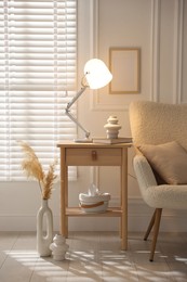 Photo of Stylish lamp, vase and book on wooden bedside table near armchair indoors