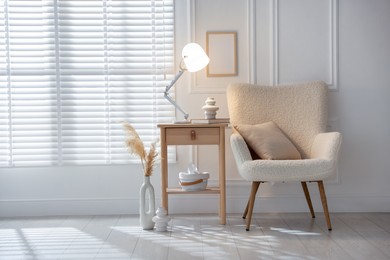 Photo of Stylish lamp, vase and book on wooden bedside table near armchair indoors, space for text