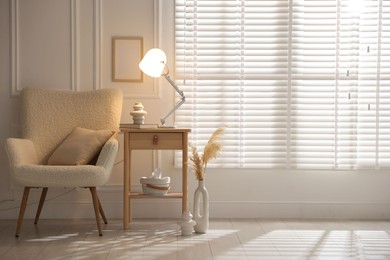 Photo of Stylish lamp, vase and book on wooden bedside table near armchair indoors, space for text