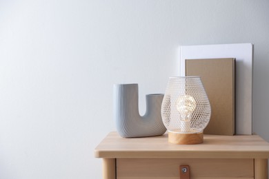 Photo of Stylish lamp, vase and books on wooden bedside table near white wall indoors, space for text