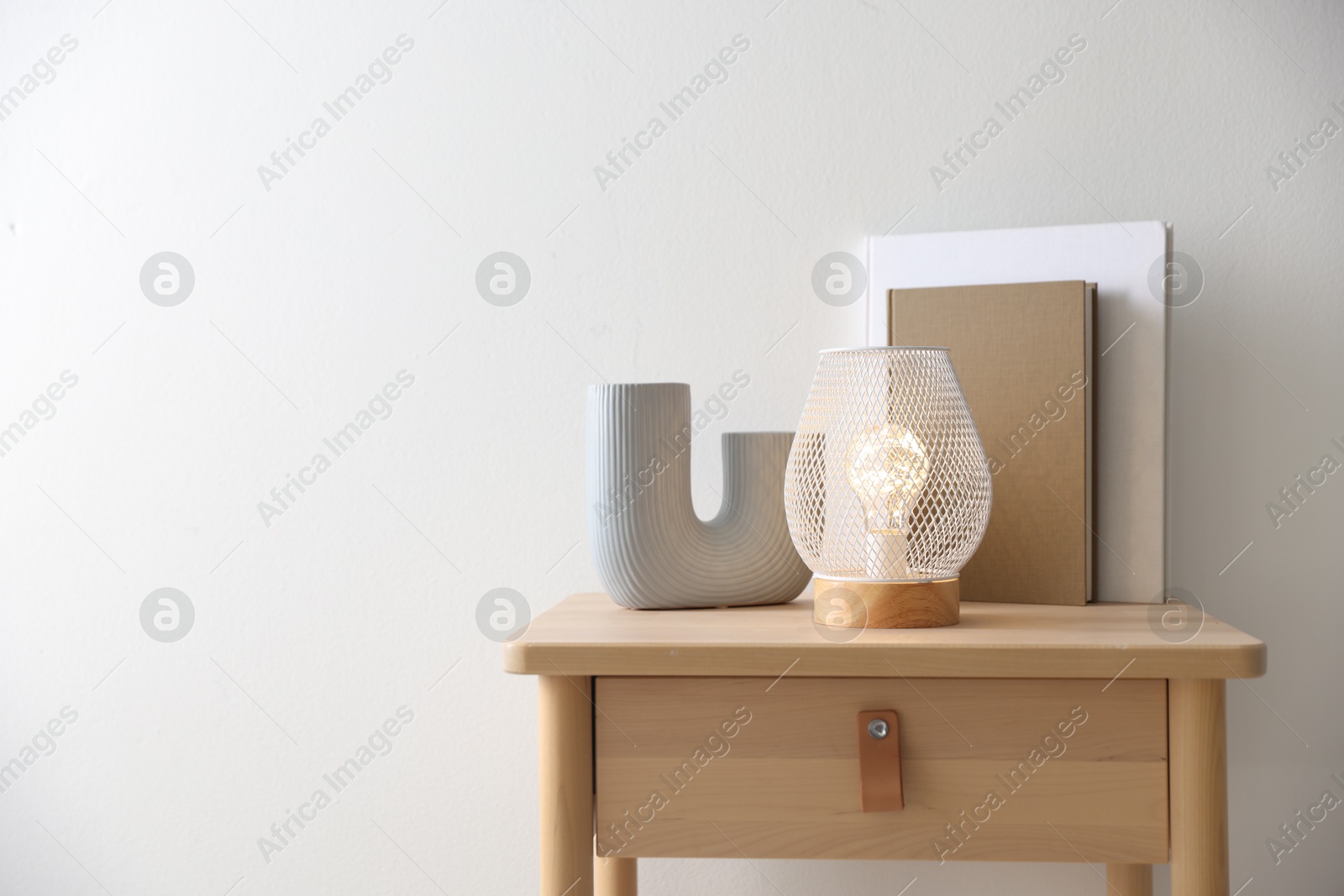 Photo of Stylish lamp, vase and books on wooden bedside table near white wall indoors, space for text