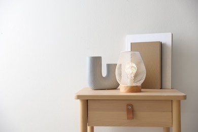 Photo of Stylish lamp, vase and books on wooden bedside table near white wall indoors, space for text