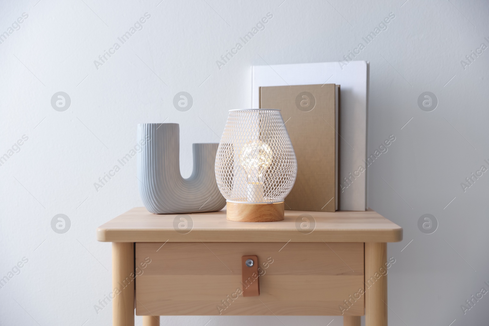 Photo of Stylish lamp, vase and books on wooden bedside table near white wall indoors
