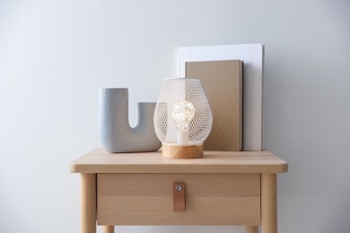 Photo of Stylish lamp, vase and books on wooden bedside table near white wall indoors