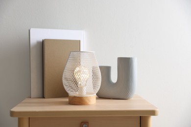 Photo of Stylish lamp, vase and books on wooden bedside table near white wall indoors