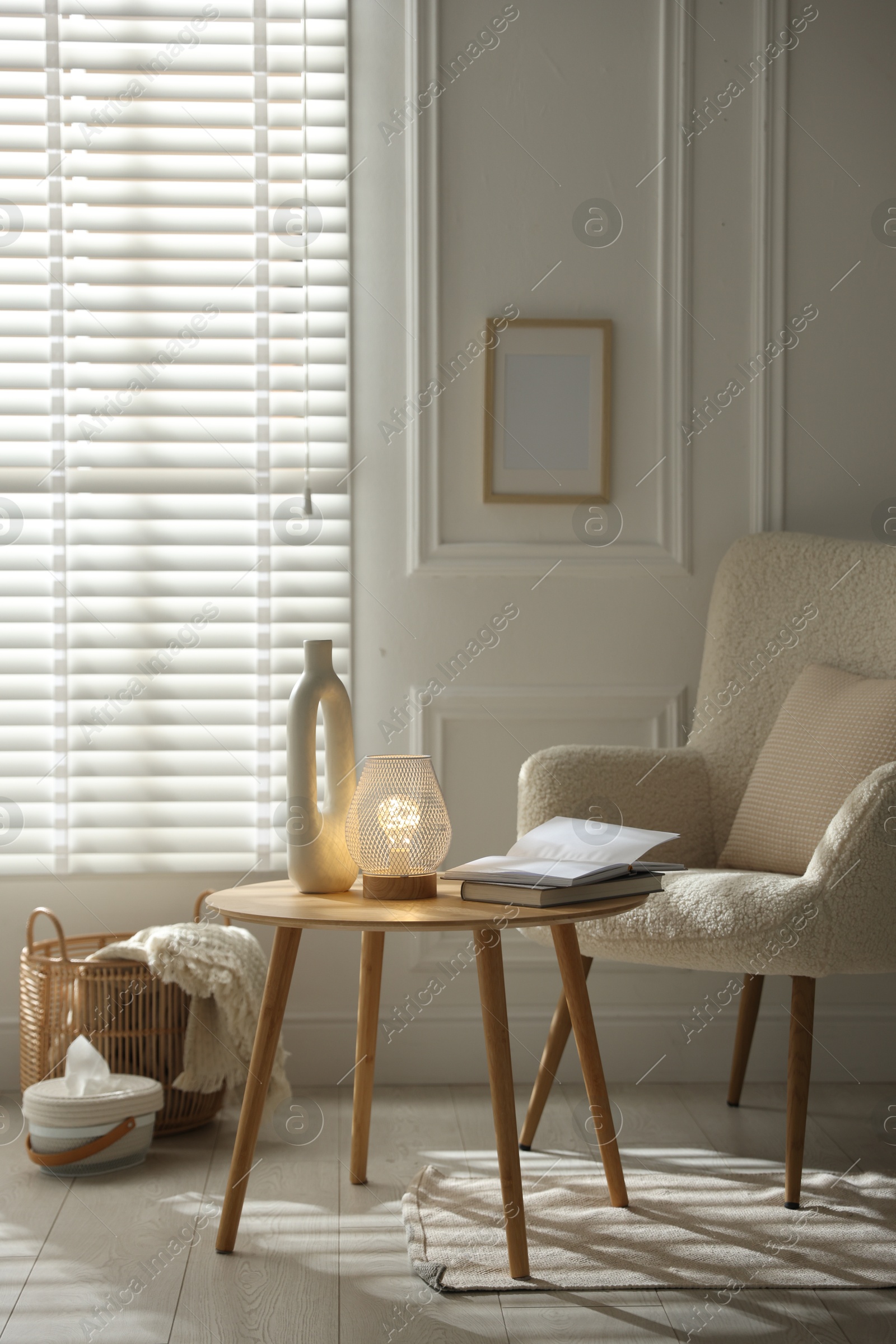 Photo of Stylish lamp, vase and books on wooden table near armchair indoors