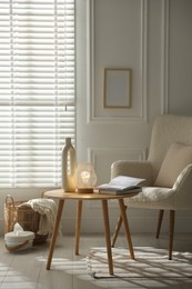 Photo of Stylish lamp, vase and books on wooden table near armchair indoors