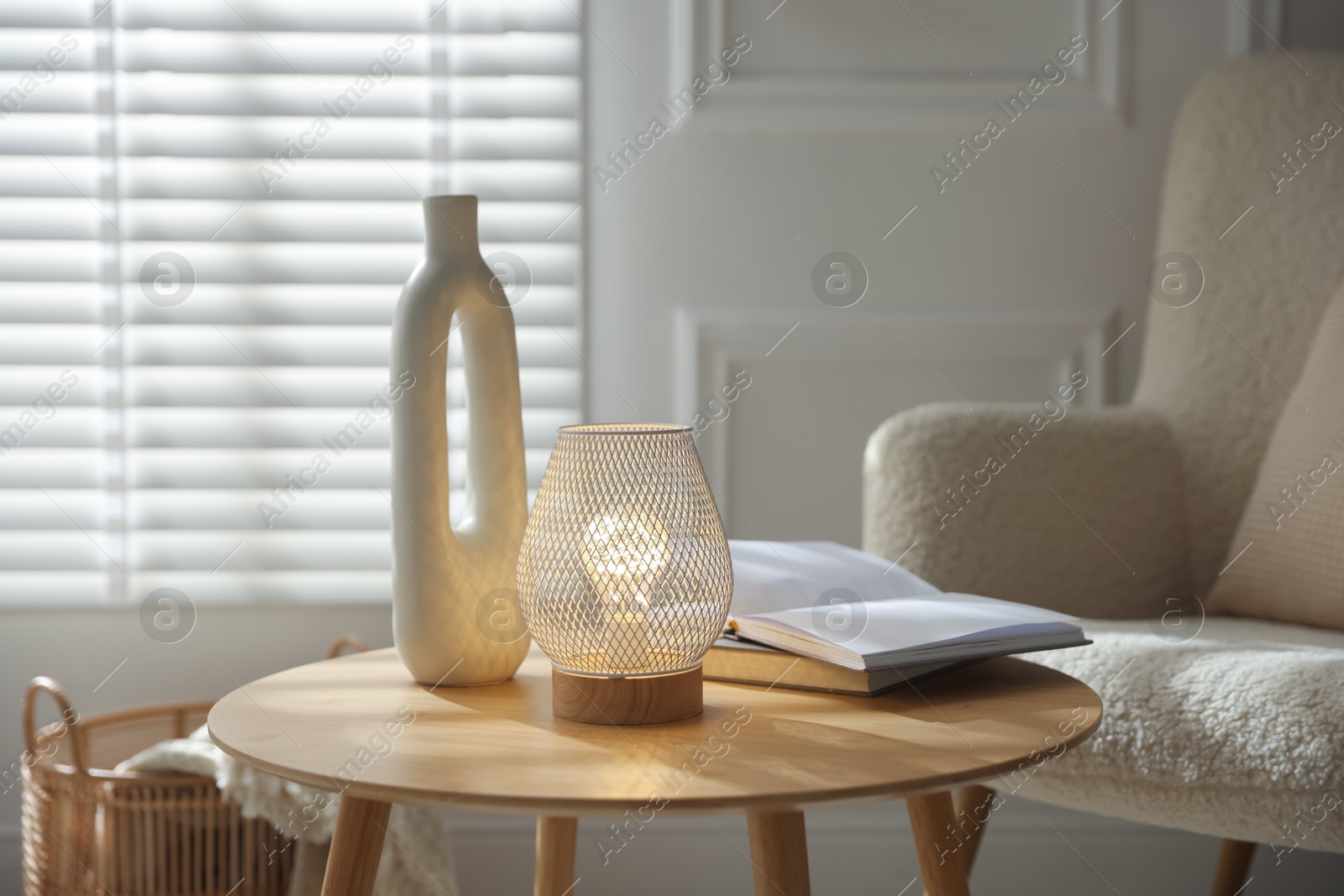 Photo of Stylish lamp, vase and books on wooden table near armchair indoors