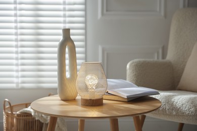 Photo of Stylish lamp, vase and books on wooden table near armchair indoors
