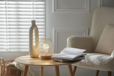 Photo of Stylish lamp, vase and books on wooden table near armchair indoors
