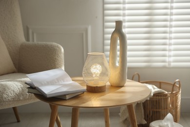 Photo of Stylish lamp, vase and books on wooden table near armchair indoors