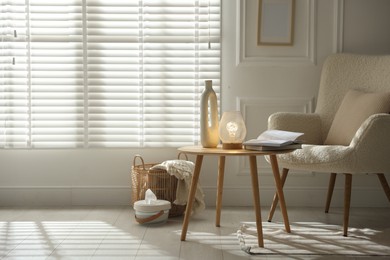 Photo of Stylish lamp, vase and books on wooden table near armchair indoors, space for text