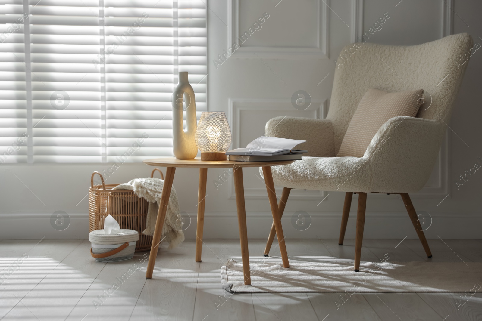 Photo of Stylish lamp, vase and books on wooden table near armchair indoors