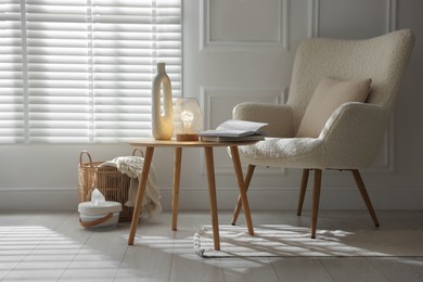 Photo of Stylish lamp, vase and books on wooden table near armchair indoors