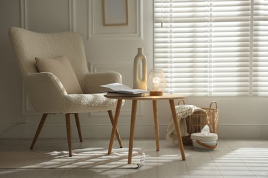 Photo of Stylish lamp, vase and books on wooden table near armchair indoors