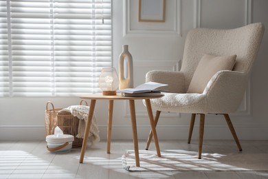 Photo of Stylish lamp, vase and books on wooden table near armchair indoors