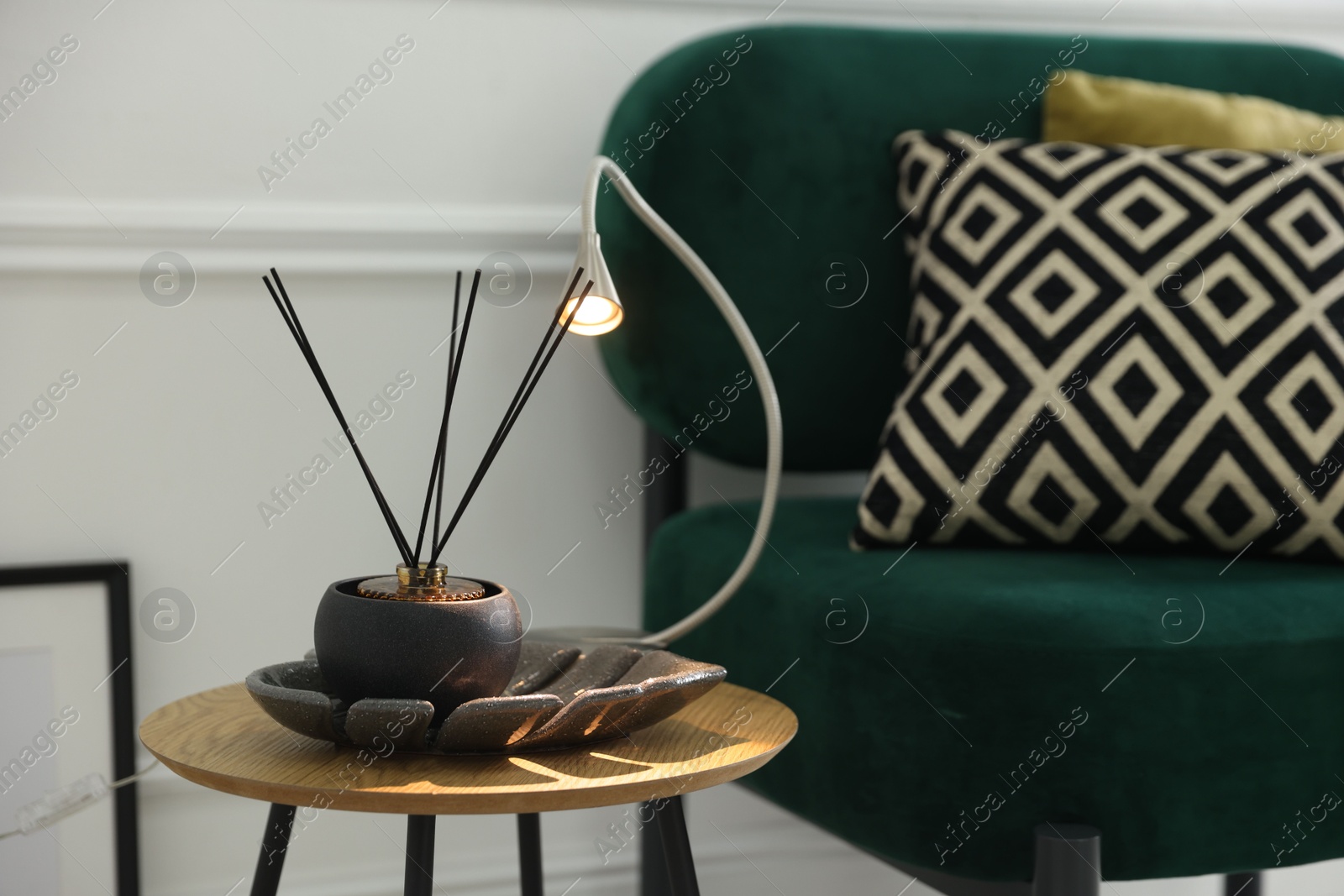 Photo of Stylish lamp and reed diffuser on side table near armchair with pillows indoors