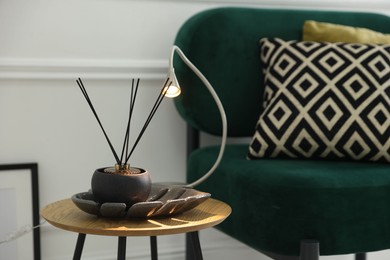 Photo of Stylish lamp and reed diffuser on side table near armchair with pillows indoors