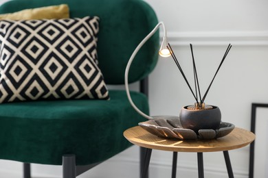 Photo of Stylish lamp and reed diffuser on side table near armchair with pillows indoors