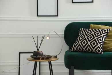 Photo of Stylish lamp and reed diffuser on side table near armchair with pillows indoors