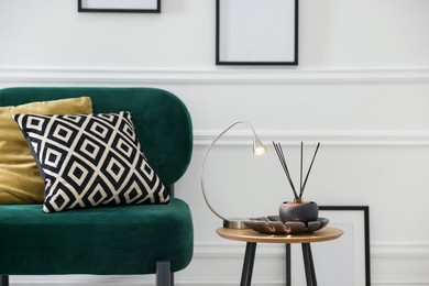 Photo of Stylish lamp and reed diffuser on side table near armchair with pillows indoors