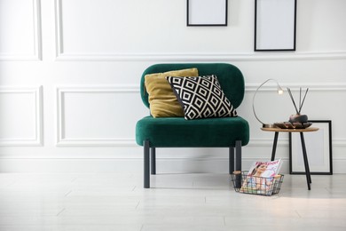 Photo of Stylish lamp, reed diffuser and magazine near armchair with pillows indoors, space for text