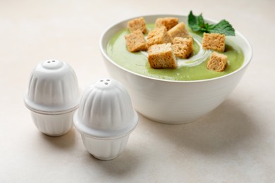 Photo of Salt and pepper shakers near bowl of delicious cream soup with croutons on light table, closeup