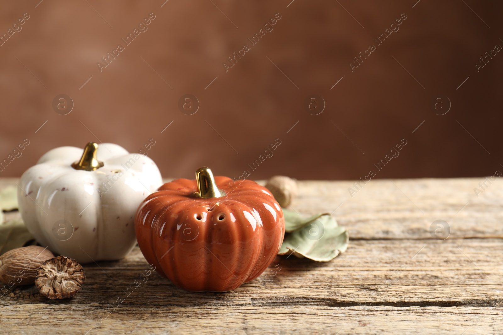 Photo of Salt, pepper shakers in shape of pumpkins, dry bay leaves and nutmegs on wooden table against blurred brown background, closeup. Space for text