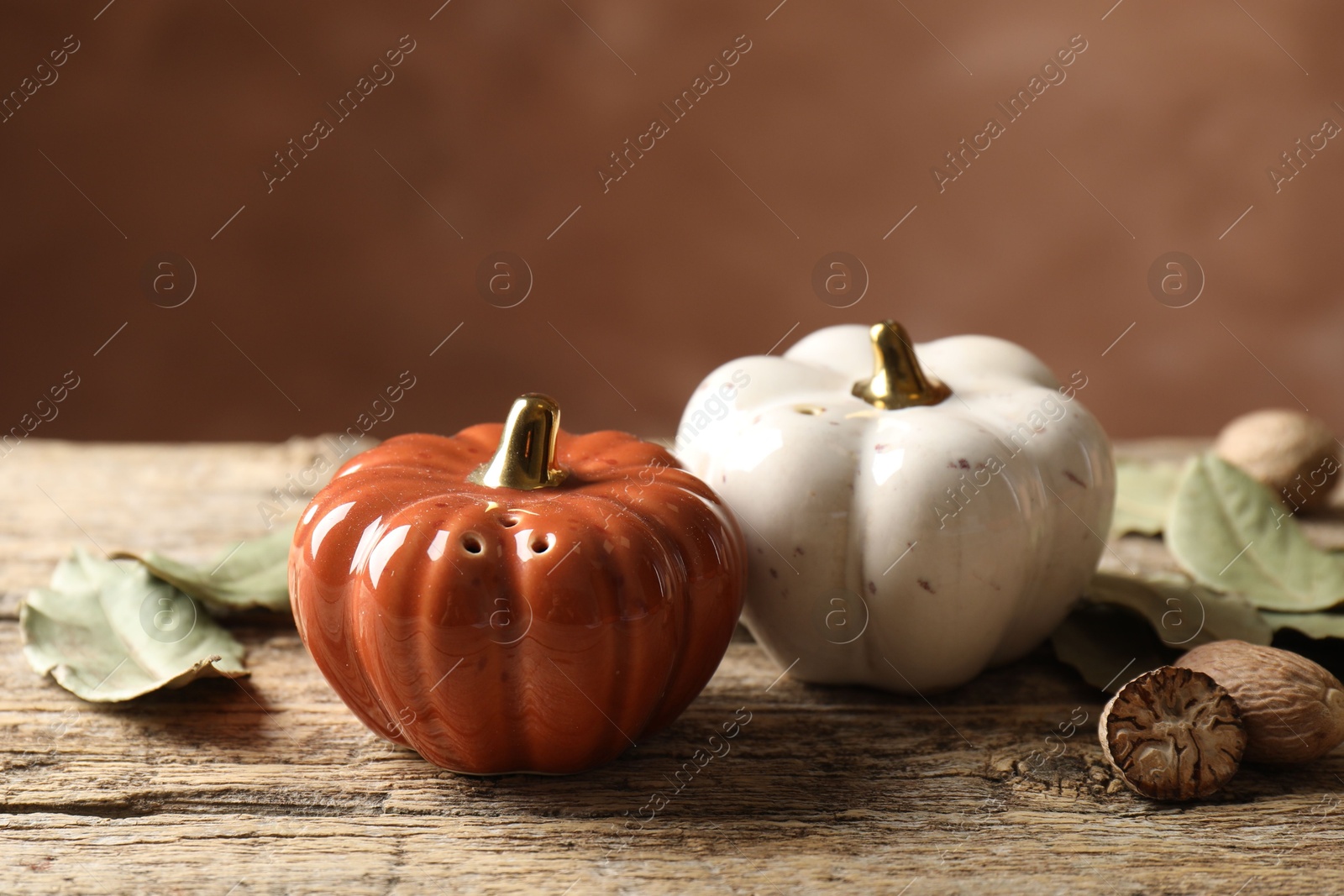 Photo of Salt, pepper shakers in shape of pumpkins, dry bay leaves and nutmegs on wooden table against blurred brown background, closeup