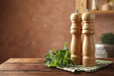 Photo of Salt, pepper mills, mint and napkin on wooden table against blurred brown background, closeup. Space for text