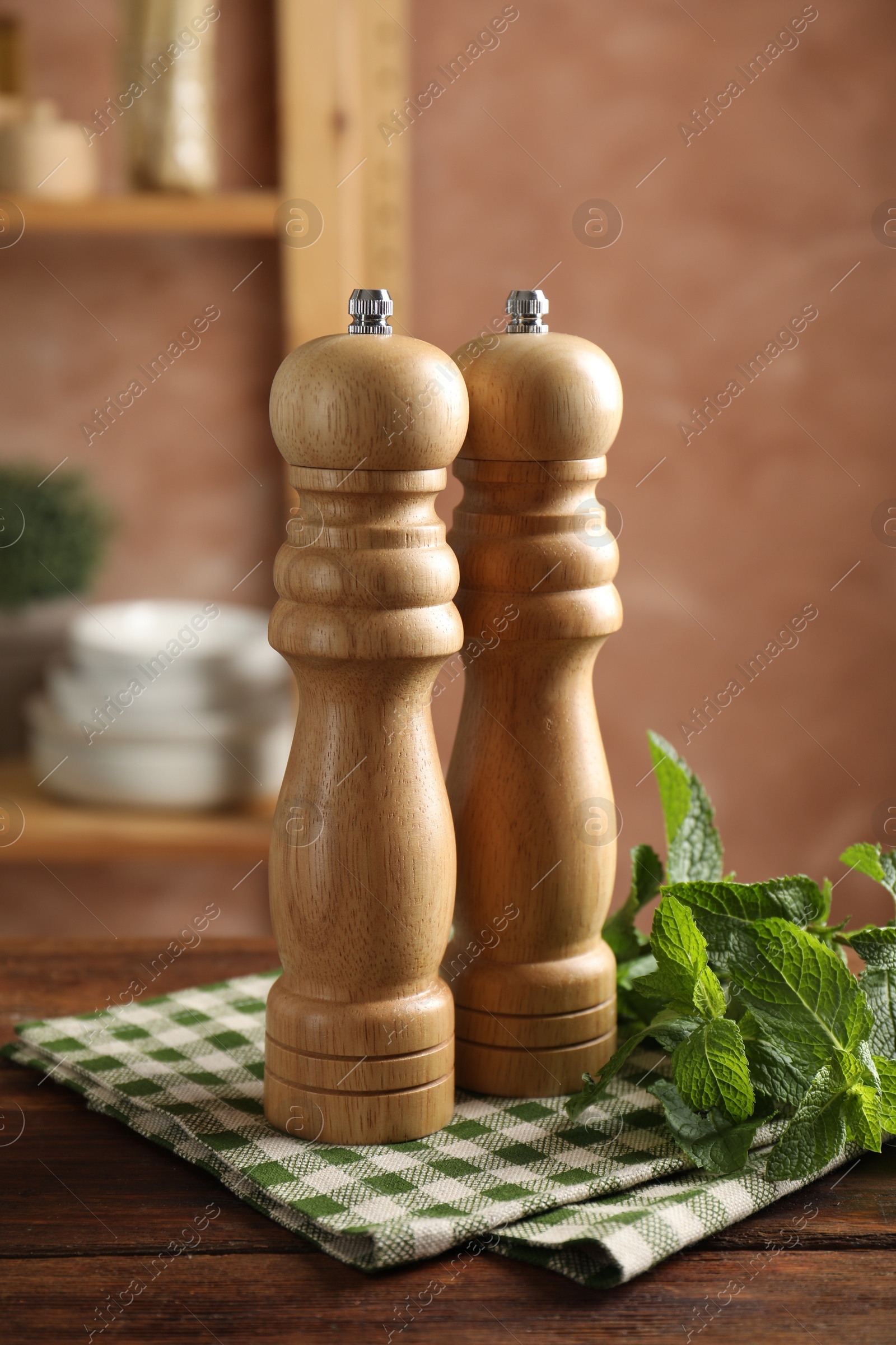 Photo of Salt, pepper mills, mint and napkin on wooden table against blurred brown background