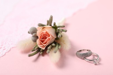 Photo of Beautiful boutonniere, wedding rings and lace fabric on light pink background, closeup