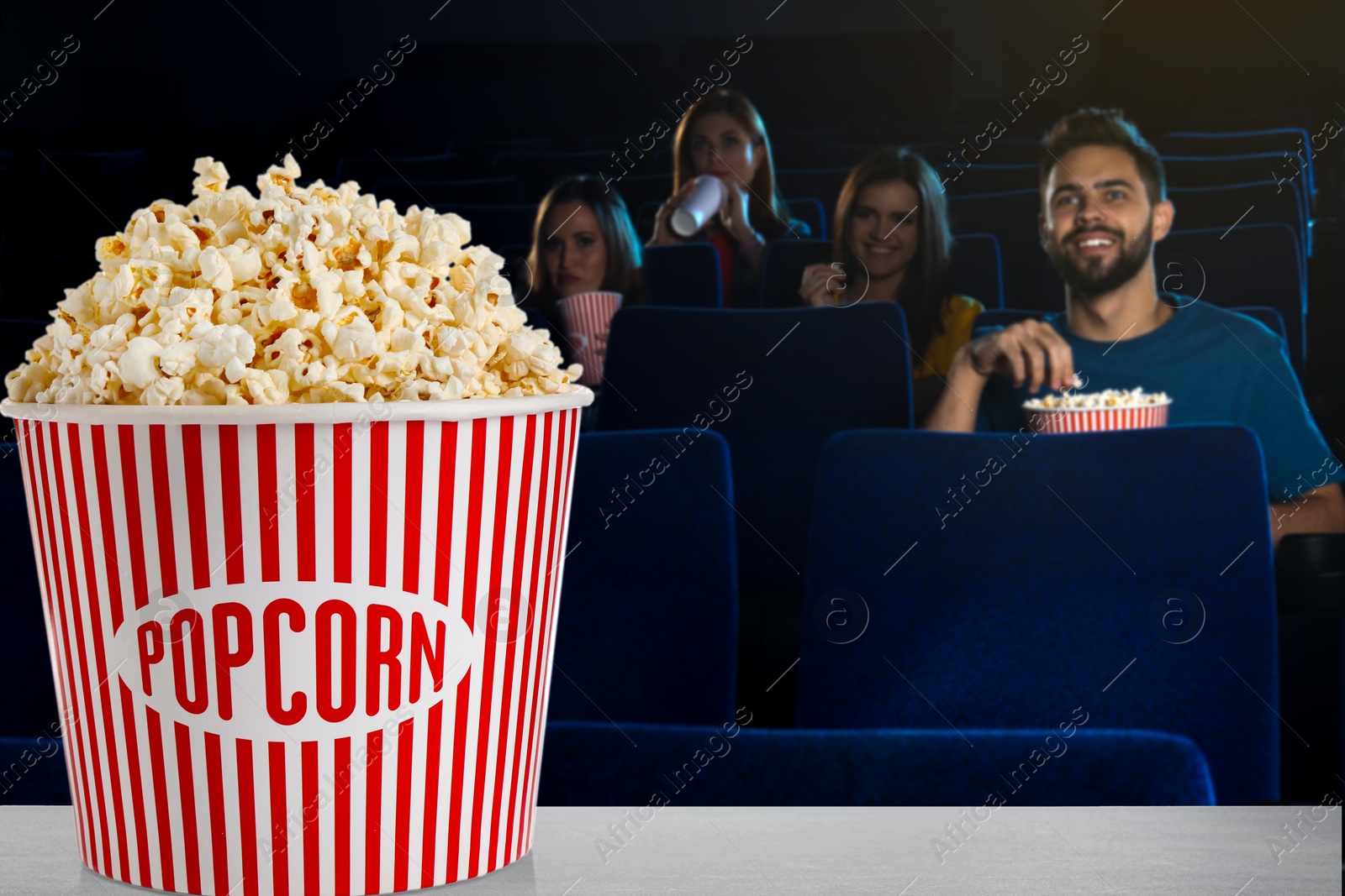 Image of Bucket of fresh popcorn on table against people watching movie in dark cinema
