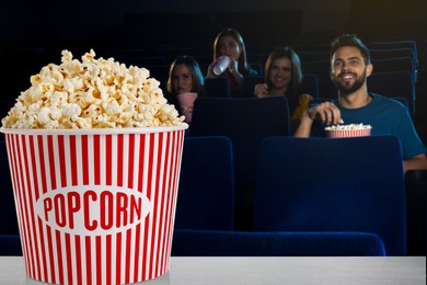 Image of Bucket of fresh popcorn on table against people watching movie in dark cinema