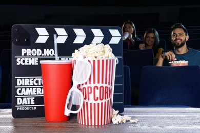 Image of Bucket of fresh popcorn, cup of drink, 3D glasses and clapper board on table against people watching movie in dark cinema