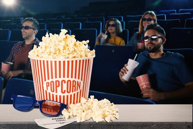 Image of Bucket of fresh popcorn, 3D glasses and tickets on table against people watching movie in dark cinema