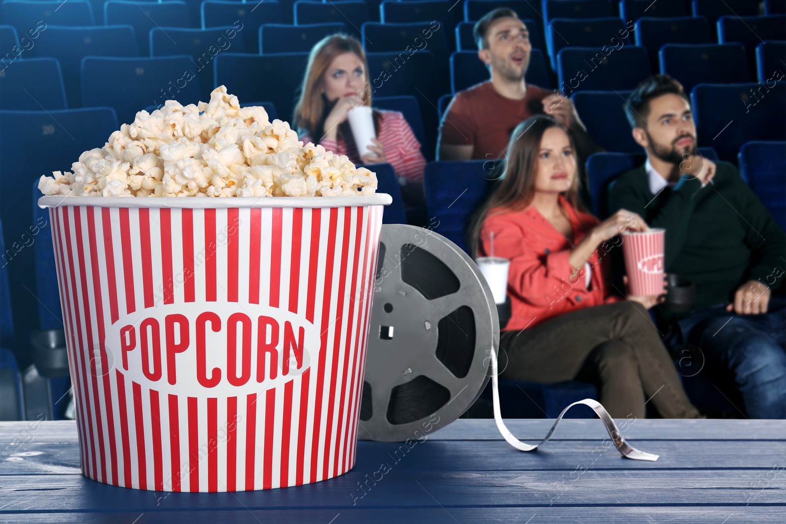 Image of Bucket of fresh popcorn and film reel on table against people watching movie in dark cinema