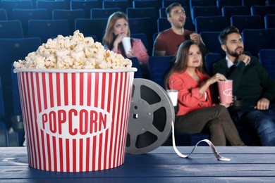 Image of Bucket of fresh popcorn and film reel on table against people watching movie in dark cinema