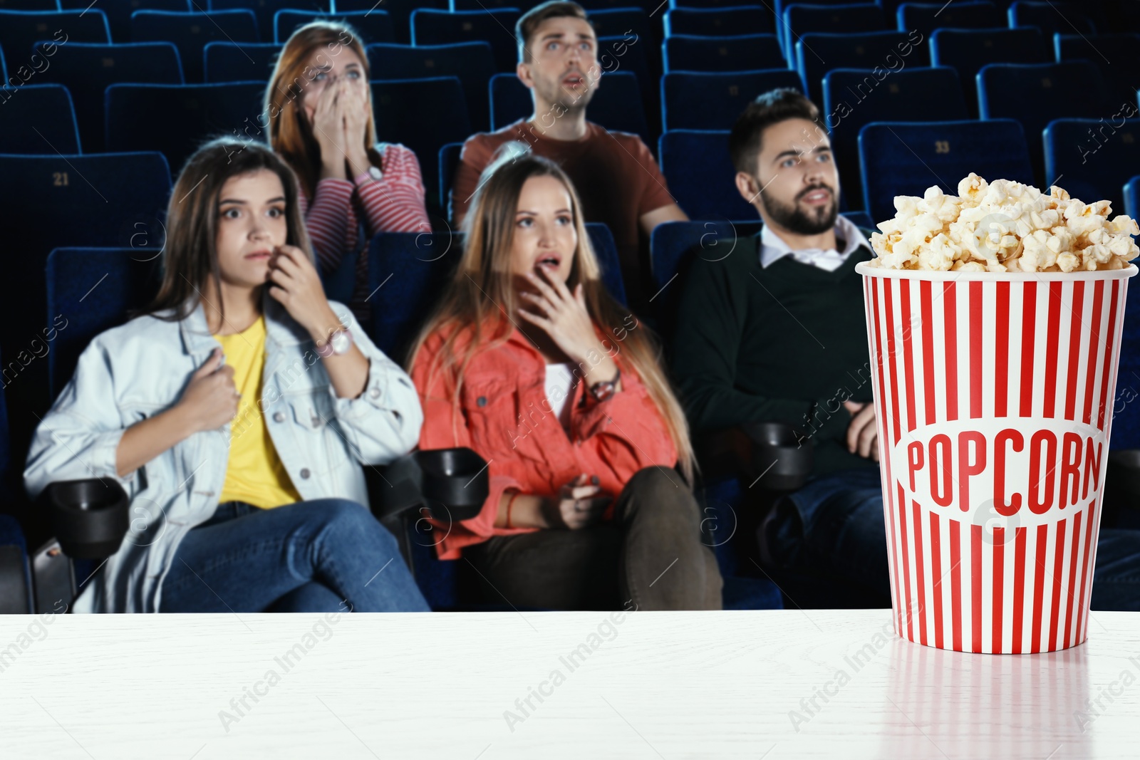 Image of Bucket of fresh popcorn on table against people watching movie in dark cinema