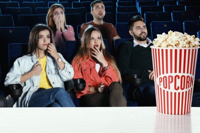 Image of Bucket of fresh popcorn on table against people watching movie in dark cinema