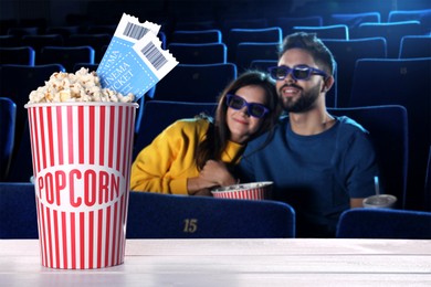 Image of Bucket of fresh popcorn and tickets on table against couple watching movie in dark cinema