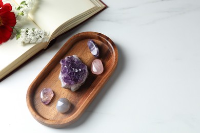 Photo of Different beautiful gemstones, book and flowers on white marble table, flat lay. Space for text
