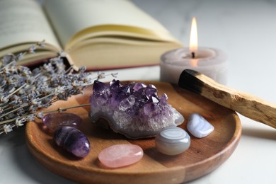 Photo of Different beautiful gemstones, palo santo, book, lavender flowers and burning candle on white table, closeup