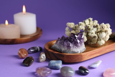 Photo of Different beautiful gemstones, flowers and burning candles on purple background, closeup
